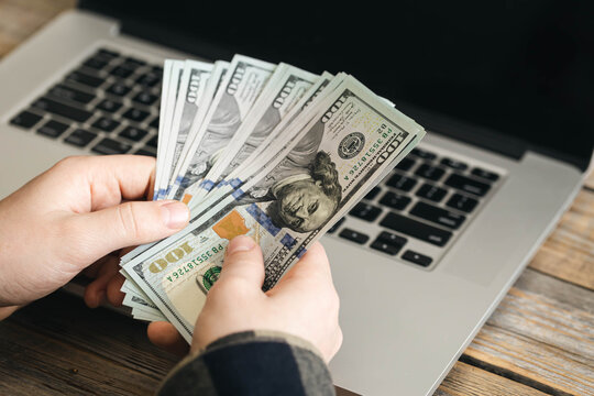 Blogging enthusiasist person Hands holding a fan of one hundred dollar bills above a laptop.