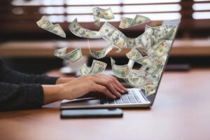 What Skills Do I Need To Make Money Online? Skills to make money online-Person typing on a laptop with hundred-dollar bills digitally flying out of the screen.